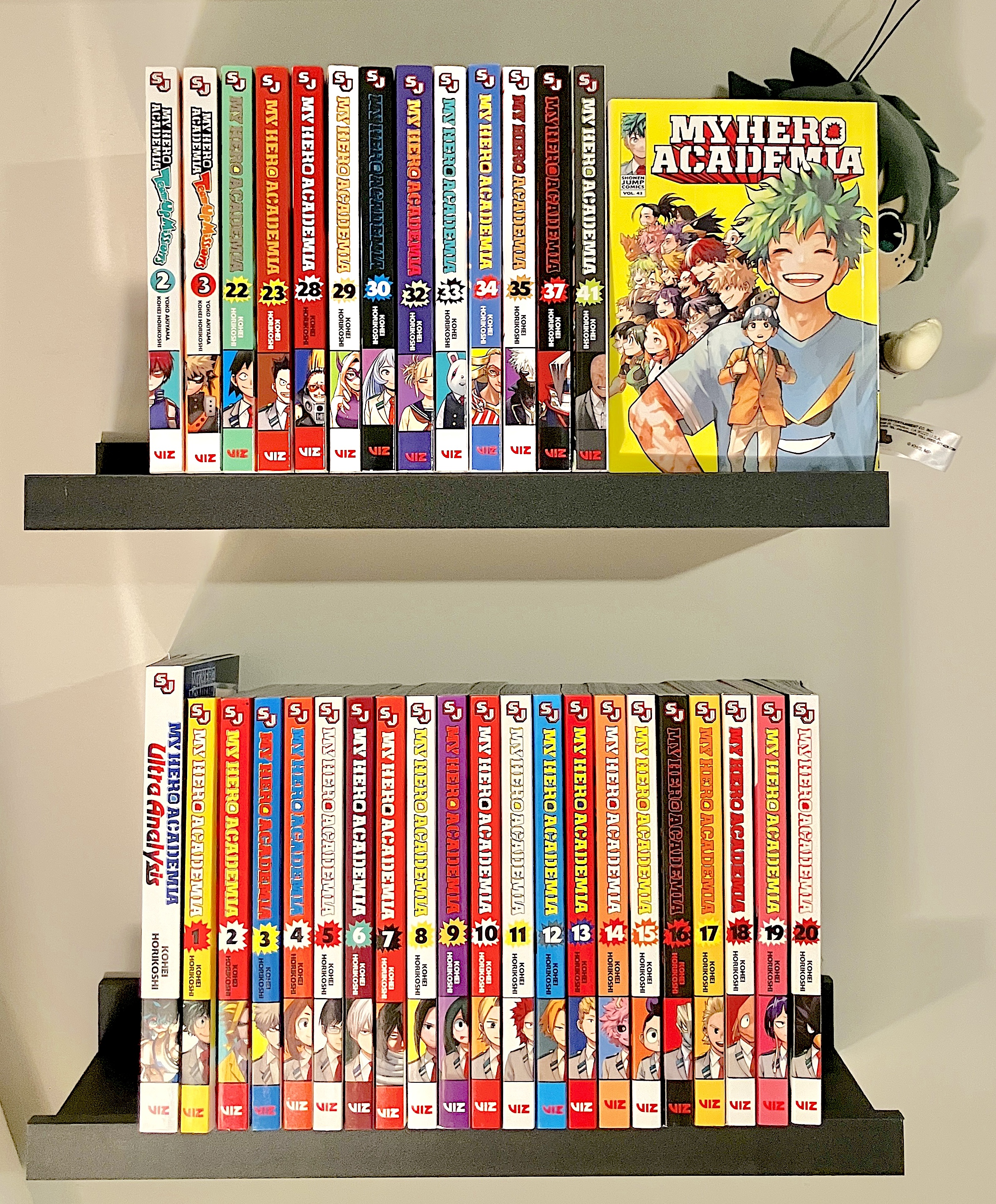 my personal shelf of mha mangas (nearly complete), with a deku plushie sitting stupidly behind them
